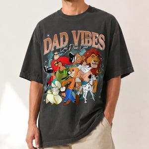Puede incluir: Camiseta gris oscuro con un dise&ntilde;o gr&aacute;fico con el texto "DAD VIBES Cool Dads Club" en naranja y blanco. El dise&ntilde;o gr&aacute;fico incluye personajes de dibujos animados como Los Incre&iacute;bles, Simba y Goofy. La camiseta es de estilo vintage.