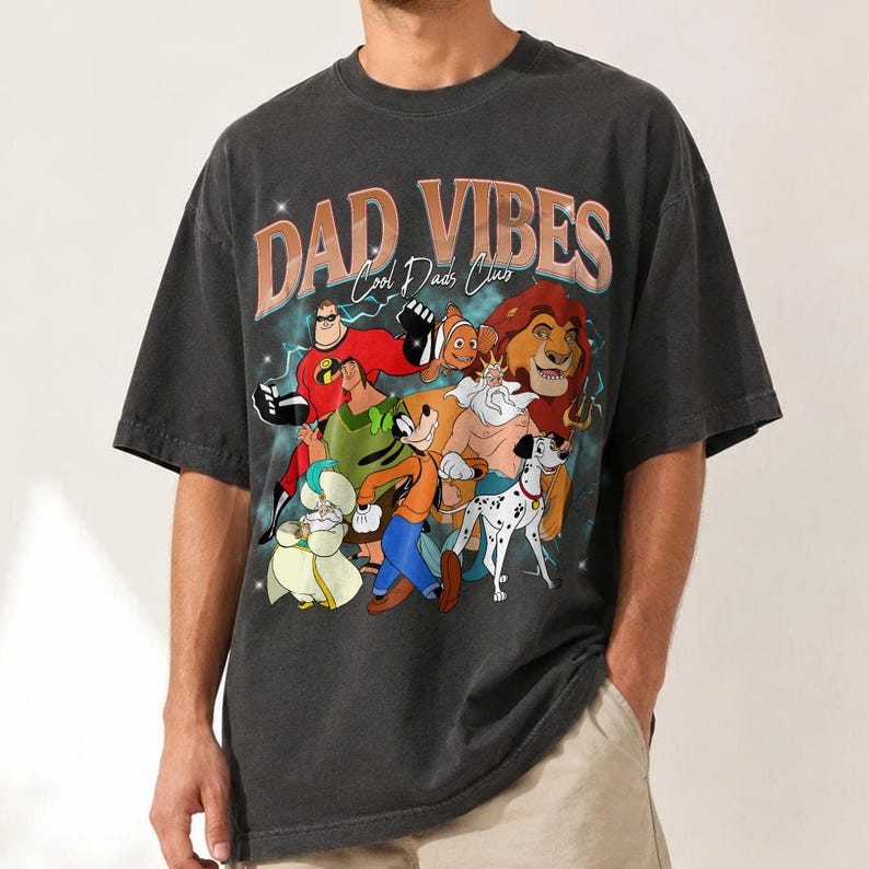 Puede incluir: Camiseta gris oscuro con un dise&ntilde;o gr&aacute;fico con el texto "DAD VIBES Cool Dads Club" en naranja y blanco. El dise&ntilde;o gr&aacute;fico incluye personajes de dibujos animados como Los Incre&iacute;bles, Simba y Goofy. La camiseta es de estilo vintage.