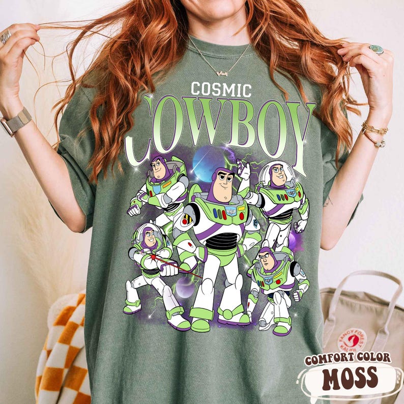 May include: A moss green t-shirt featuring the text "COSMIC COWBOY" in a light green font above a graphic illustration of five Buzz Lightyear characters in various poses. The design includes purple and blue cosmic elements and lightning effects.