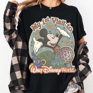 May include: Black t-shirt featuring a graphic of Mickey Mouse with the text "My 1st Visit to Walt Disney World." The design includes a castle, a sorcerer's hat, and a geodesic sphere. The shirt has a vintage, retro style.