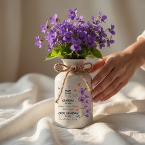 May include: A white ceramic vase with a bouquet of purple flowers, tied with a jute bow. The vase is decorated with floral and butterfly designs, and text that reads "MOM Est. 1971", "GRANDMA Est. 2000", and "GREAT GRANDMA Est. 2005".