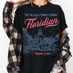 May include: Black t-shirt featuring the text "The Villas at Disney's Grand Floridian Resort & Spa" in pink and blue. The design includes an illustration of the resort. The shirt is a casual wear item.