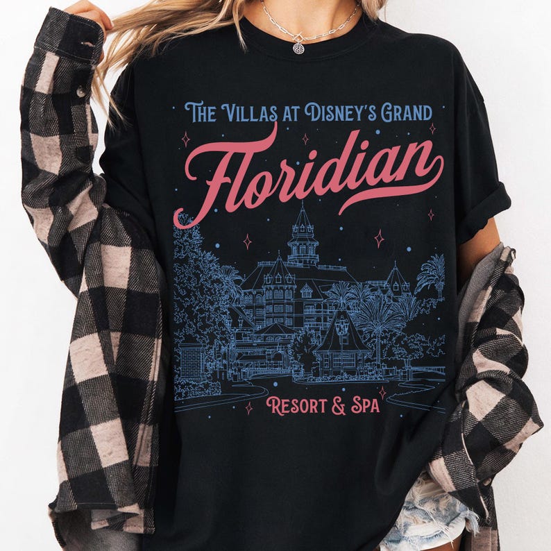 May include: Black t-shirt featuring the text "The Villas at Disney's Grand Floridian Resort & Spa" in pink and blue. The design includes an illustration of the resort. The shirt is a casual wear item.