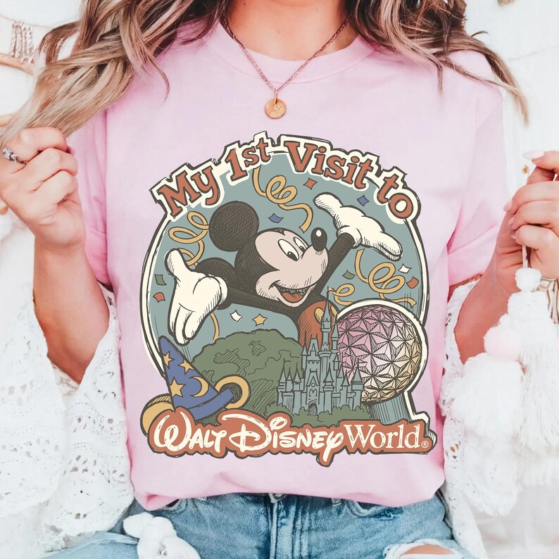 May include: A light pink t-shirt featuring a vintage-style graphic of Mickey Mouse with the text "My 1st Visit to Walt Disney World." The design includes a castle, a wizard's hat, and a geometric sphere.