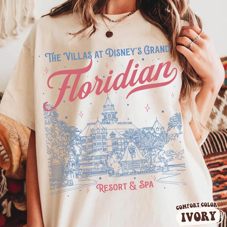 May include: Ivory-colored t-shirt featuring the text "The Villas at Disney's Grand Floridian Resort & Spa" in pink and blue. The design includes an illustration of the resort. The shirt is a comfort color.