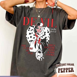 May include: Dark gray t-shirt featuring a Cruella De Vil graphic. The design includes the text "DEVIL" in red, the character's name, and the phrases "THE FUTURE OF FUR" and "SHE'S GOT THE WITS & THE LOOKS". The shirt is labeled "COMFORT COLOR PEPPER".