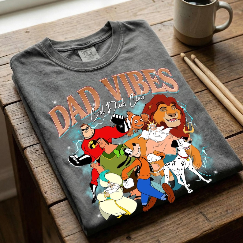 Puede incluir: Camiseta gris con las palabras "DAD VIBES" y "Cool Dads Club" en color cobre. La camiseta presenta personajes de dibujos animados como Mr. Incre&iacute;ble, Simba y Goofy. La camiseta est&aacute; doblada sobre una superficie de madera.