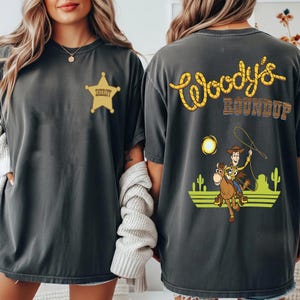 K&ouml;nnte beinhalten: Dunkelgraue T-Shirts mit einem Sheriff-Abzeichen und "Woody's Roundup"-Grafik. Auf der R&uuml;ckseite des Shirts ist ein Cartoon-Cowboy zu sehen, der mit einem Lasso auf einem Pferd reitet, vor einer W&uuml;stenlandschaft mit Kakteen und einer Sonne.
