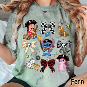 Może przedstawiać: Jasnozielona koszulka z r&oacute;żnymi postaciami pirat&oacute;w z kresk&oacute;wek i wzorami kokard. Koszulka zawiera wizerunki pirata grającego na gitarze, niebieskiego kosmity trzymającego pochodnię i r&oacute;żowego kosmity w pirackim kapeluszu.