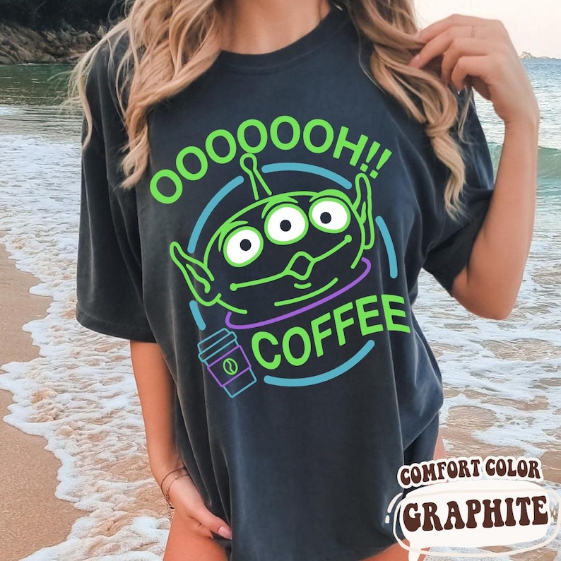 Puede incluir: Camiseta gris oscuro con un dise&ntilde;o de alien&iacute;gena verde y el texto "OOOOOOH!! COFFEE". El dise&ntilde;o incluye una taza de caf&eacute; y est&aacute; delineado en verde, morado y azul. La camiseta est&aacute; etiquetada como "COMFORT COLOR GRAPHITE".