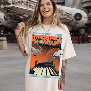 May include: Cream-colored t-shirt featuring a graphic design for Star Wars Hyperspace Mountain at Disneyland. The design includes spaceships, a mountain silhouette, and the text "Star Wars Hyperspace Mountain" in orange and blue.
