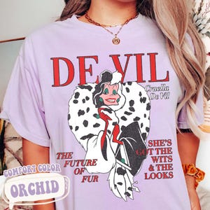 May include: Orchid-colored t-shirt featuring a graphic of Cruella De Vil with the text "DE VIL" in red. The shirt also includes the phrases "THE FUTURE OF FUR" and "SHE'S GOT THE WITS & THE LOOKS."