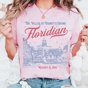 May include: Pink t-shirt with the text "The Villas at Disney's Grand Floridian Resort & Spa" in red and blue. The design includes an illustration of the resort. The shirt is a light pink color.