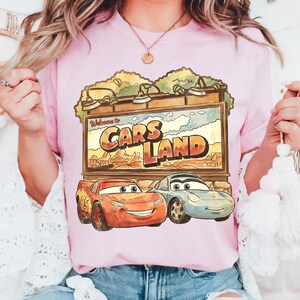 Op de afbeelding: Lichtroze T-shirt met een Cars Land-afbeelding met Bliksem McQueen en Sally Carrera. Het bord leest "Welcome to Cars Land" tegen een woestijnachtergrond. Het shirt heeft een lichtroze kleur.