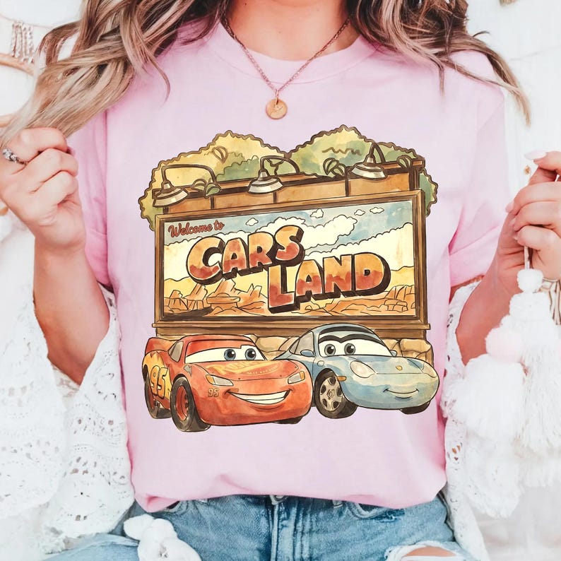 Op de afbeelding: Lichtroze T-shirt met een Cars Land-afbeelding met Bliksem McQueen en Sally Carrera. Het bord leest "Welcome to Cars Land" tegen een woestijnachtergrond. Het shirt heeft een lichtroze kleur.