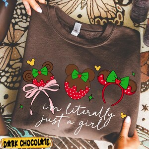 May include: Dark chocolate brown sweatshirt with a graphic of three Mickey Mouse-inspired designs. The designs include a bow, a strawberry, and the text "I'm literally just a girl." The sweatshirt has a crew neck.