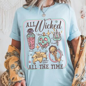 May include: Light blue t-shirt with a colorful graphic design. The design features the words "ALL Wicked ALL THE TIME" with illustrations of a poison bottle, skull, trident, and other elements. The shirt is a short-sleeved crew neck.