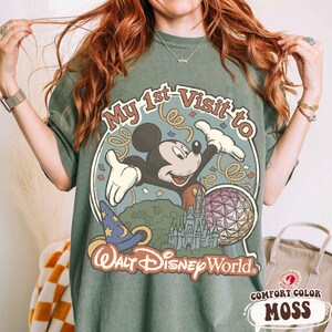 May include: Moss green t-shirt featuring a graphic of Mickey Mouse with the text "My 1st Visit to Walt Disney World." The design includes a castle, a wizard hat, and a colorful sphere. The shirt is a comfort color.