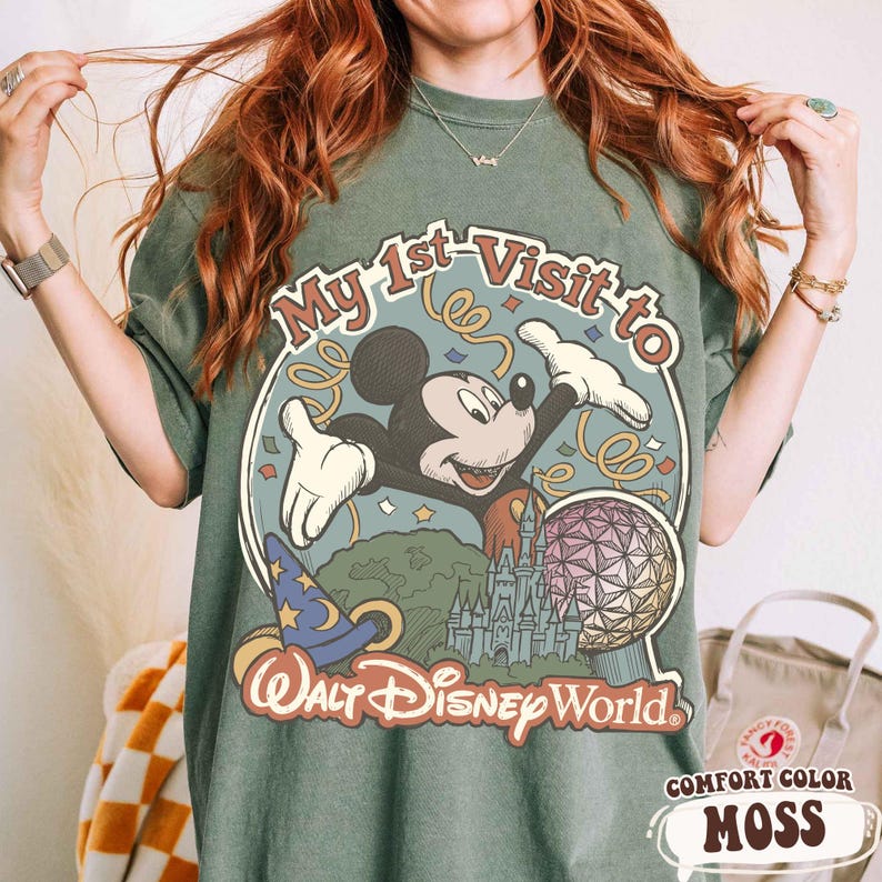 May include: Moss green t-shirt featuring a graphic of Mickey Mouse with the text "My 1st Visit to Walt Disney World." The design includes a castle, a wizard hat, and a colorful sphere. The shirt is a comfort color.