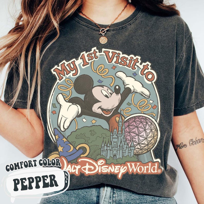 May include: Pepper-colored t-shirt with a graphic featuring Mickey Mouse and the text "My 1st Visit to Walt Disney World." The design includes a castle, a sphere, and a wizard's hat. The shirt is labeled "Comfort Color."