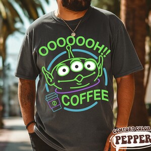 Puede incluir: Camiseta gris oscuro con un gr&aacute;fico de alien&iacute;gena verde y el texto "OOOOOOH!! COFFEE". El dise&ntilde;o incluye un icono de taza de caf&eacute; y est&aacute; rodeado de l&iacute;neas azules y moradas. La camiseta est&aacute; etiquetada como "COMFORT COLOR PEPPER".