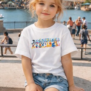 Puede incluir: Una camiseta blanca con un gr&aacute;fico colorido de personajes de Disney sosteniendo carteles que deletrean "Disneyland". La camiseta la lleva una ni&ntilde;a peque&ntilde;a que lleva pantalones cortos de mezclilla. El fondo muestra una escena frente al mar con gente.