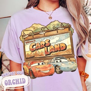 Op de afbeelding: Orchidee-kleurig T-shirt met een afbeelding van Cars Land met Bliksem McQueen en Sally Carrera. Het ontwerp bevat de tekst "Welcome to Cars Land" en "Comfort Color".