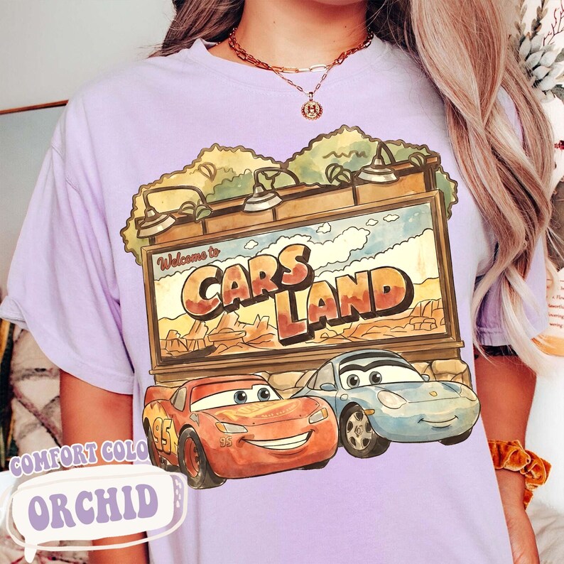 Op de afbeelding: Orchidee-kleurig T-shirt met een afbeelding van Cars Land met Bliksem McQueen en Sally Carrera. Het ontwerp bevat de tekst "Welcome to Cars Land" en "Comfort Color".