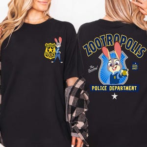 May include: Black t-shirt featuring a Zootopia Police Department design. The front displays a badge with Judy Hopps, while the back showcases the Zootropolis logo and Judy Hopps. The shirt includes the text "New Recruiting!" and "Keeping Critters Safe!"