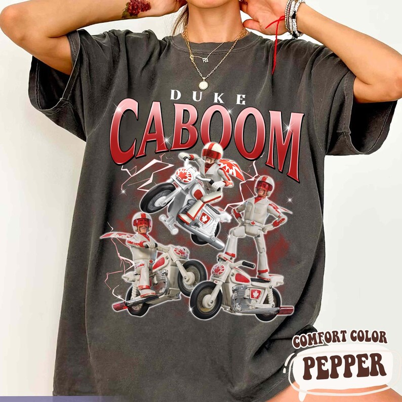 May include: Dark grey t-shirt featuring the text "DUKE CABOOM" in red, with a graphic of a character on a motorcycle. The graphic includes three figures on white and red motorcycles. The shirt is labeled "COMFORT COLOR PEPPER".