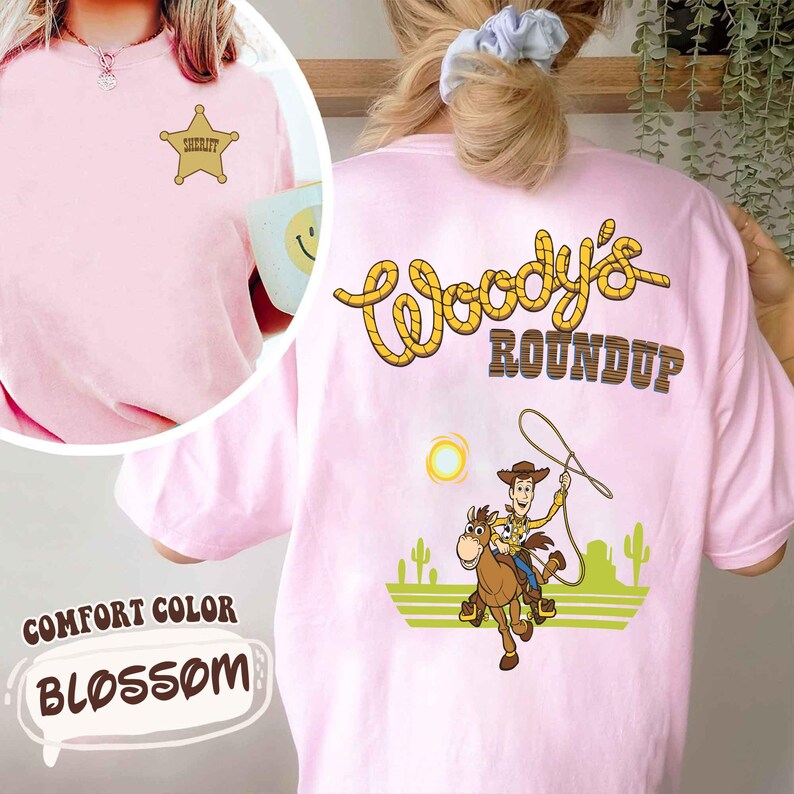 K&ouml;nnte beinhalten: Hellrosa T-Shirt mit dem Text "Woody's Roundup" in Gelb und Braun. Auf der R&uuml;ckseite des Shirts ist ein Cartoon-Cowboy auf einem Pferd abgebildet. Auf der Vorderseite befindet sich ein Sheriff-Abzeichen. Das Shirt ist mit "Comfort Color Blossom" gekennzeichnet.