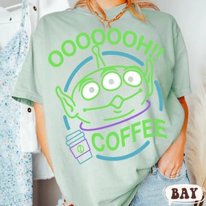 Puede incluir: Camiseta verde claro con un dise&ntilde;o de extraterrestre de dibujos animados con tres ojos y el texto "OOOOOOH!! COFFEE" en verde y morado. El extraterrestre est&aacute; dentro de un c&iacute;rculo azul, y tambi&eacute;n se muestra una taza de caf&eacute;.