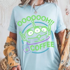 Puede incluir: Camiseta azul claro con un gr&aacute;fico de alien&iacute;gena verde y el texto "OOOOOOH!! COFFEE". El alien&iacute;gena tiene tres ojos y orejas grandes. Tambi&eacute;n se incluye una taza de caf&eacute; morada en el dise&ntilde;o.