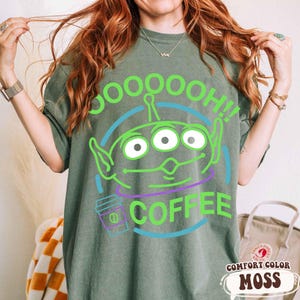 Puede incluir: Camiseta verde con un gr&aacute;fico de un alien&iacute;gena de tres ojos y las palabras "SOOOOOOH!! COFFEE". El alien&iacute;gena es verde con acentos azules y morados. La camiseta es de la marca Comfort Color en color Moss.