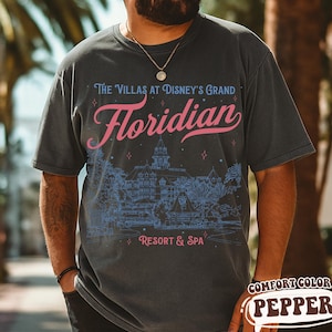 May include: Dark gray t-shirt with a graphic design featuring the text "The Villas at Disney's Grand Floridian Resort & Spa" in pink and blue. The design includes an illustration of the resort. The shirt is a Comfort Color Pepper.