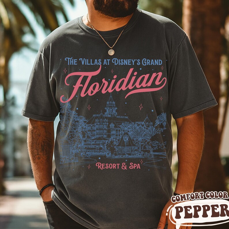 May include: Dark gray t-shirt with a graphic design featuring the text "The Villas at Disney's Grand Floridian Resort & Spa" in pink and blue. The design includes an illustration of the resort. The shirt is a Comfort Color Pepper.