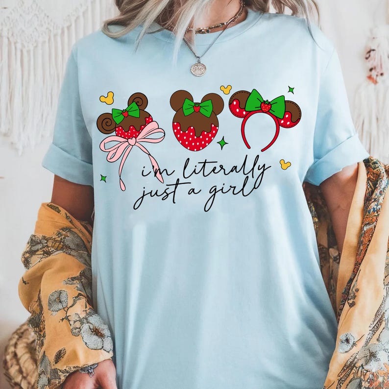 May include: Light blue t-shirt featuring three Mickey Mouse-inspired designs with bows and strawberries. The text "I'm literally just a girl" is written in a script font. The shirt has rolled-up sleeves.