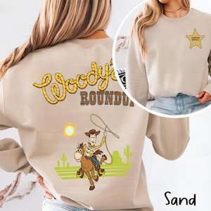 K&ouml;nnte beinhalten: Sandfarbenes Sweatshirt mit einem Cartoon-Cowboy-Motiv und dem Text "Woody's Roundup" in Gelb und Braun. Das Design umfasst einen Cowboy auf einem Pferd, einen Lasso und ein Sheriff-Abzeichen.