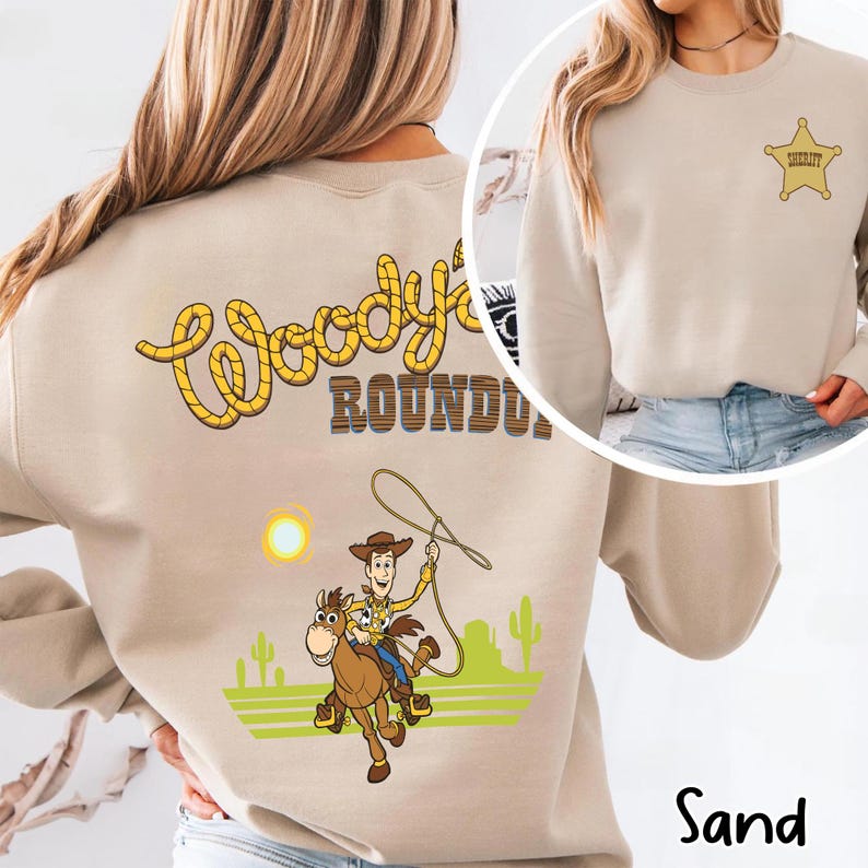 K&ouml;nnte beinhalten: Sandfarbenes Sweatshirt mit einem Cartoon-Cowboy-Motiv und dem Text "Woody's Roundup" in Gelb und Braun. Das Design umfasst einen Cowboy auf einem Pferd, einen Lasso und ein Sheriff-Abzeichen.