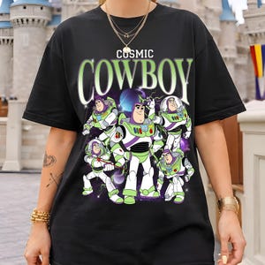 May include: A black short-sleeve t-shirt featuring a graphic print of multiple Buzz Lightyear characters in various poses against a cosmic background. The text "COSMIC COWBOY" is printed in large, light green letters above the character illustrations.