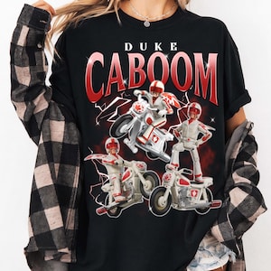 May include: Black t-shirt featuring the text "DUKE CABOOM" in red, with a graphic of a toy character on a motorcycle. The character is wearing a red helmet and white outfit. Lightning bolt graphics are in the background.