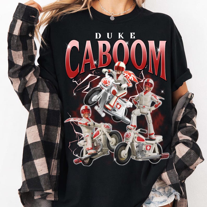 May include: Black t-shirt featuring the text "DUKE CABOOM" in red, with a graphic of a toy character on a motorcycle. The character is wearing a red helmet and white outfit. Lightning bolt graphics are in the background.