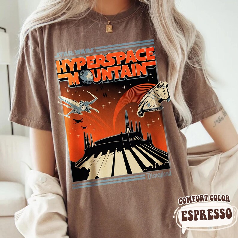 May include: Brown t-shirt featuring a retro-style graphic for Star Wars Hyperspace Mountain at Disneyland. The design includes an X-wing fighter, the Millennium Falcon, and the ride's silhouette against an orange and black background. The shirt is labeled "Comfort Color Espresso."