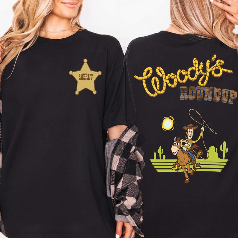 K&ouml;nnte beinhalten: Schwarzes T-Shirt mit einem goldenen Sheriff-Abzeichen auf der Vorderseite. Auf der R&uuml;ckseite des Shirts steht der Text "Woody's Roundup" in Gelb und Blau sowie ein Cartoon-Bild von Woody auf einem Pferd.