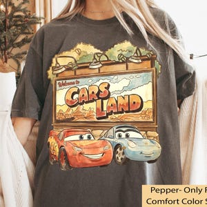 Op de afbeelding: Donkergrijs t-shirt met een kleurrijke afbeelding van het Cars Land-bord met Bliksem McQueen en Sally Carrera. Het bord leest "Welcome to Cars Land" in oranje en rode letters.