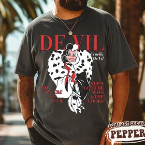 May include: Dark grey t-shirt featuring a graphic of Cruella De Vil with the text "DEVIL" in red. The design includes the phrases "THE FUTURE OF FUR" and "SHE'S GOT THE WITS & THE LOOKS". The shirt is labeled "COMFORT COLOR PEPPER".