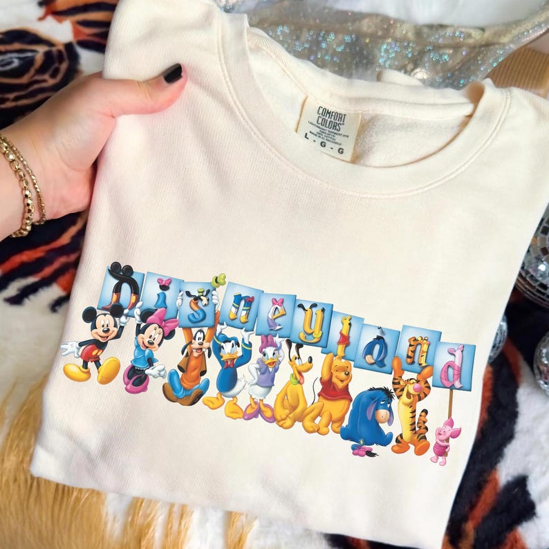 Puede incluir: Sudadera color crema con un gr&aacute;fico de personajes de Disney sosteniendo letras que deletrean "Disneyland". Los personajes incluyen Mickey Mouse, Minnie Mouse, Donald Duck y Winnie the Pooh. La sudadera tiene una etiqueta Comfort Colors.
