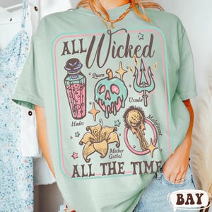 May include: A light green t-shirt with a graphic design featuring the text "ALL Wicked ALL THE TIME." The design includes illustrations of a poison bottle, a skull, a trident, a flower, and a magic mirror. The names Hades, Queen, Ursula, Maleficent, and Mother Gothel are also included.
