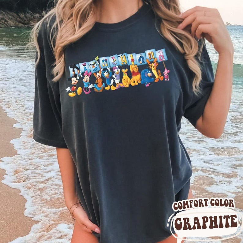 Puede incluir: Camiseta de color grafito con un gr&aacute;fico colorido de personajes de Disney, incluyendo Mickey Mouse y Winnie the Pooh. La camiseta tiene las palabras "Disneyland" encima de los personajes y "Comfort Color Graphite" debajo.