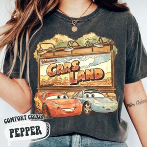 Op de afbeelding: Donkergrijs t-shirt met een kleurrijke afbeelding van het Cars Land-bord met Bliksem McQueen en Sally Carrera. Op het shirt staan de woorden "Welcome to Cars Land" en "Comfort Color Pepper".
